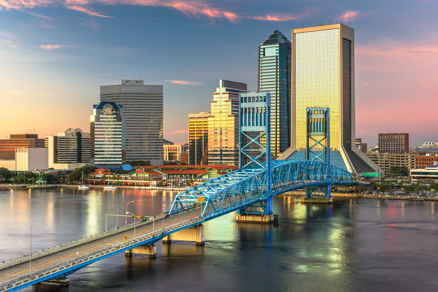 Downtown Jacksonville, Florida skyline at sunset, features modern skyscrapers and the blue Main Street Bridge spanning the St. Johns River, with reflections on the water.