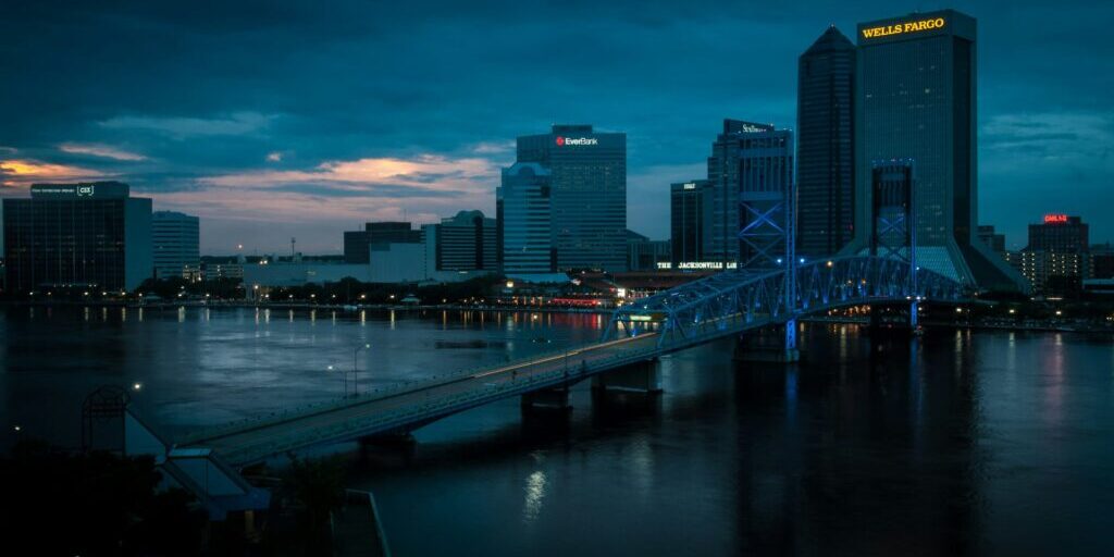 A city skyline at dusk features tall office buildings with illuminated signs and a blue-lit bridge spanning a calm river under a partly cloudy sky with hints of sunset.