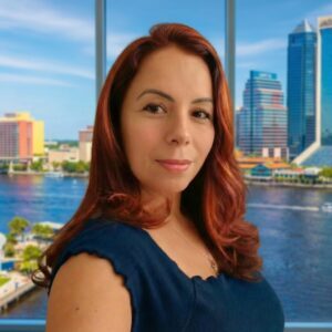 A woman with long red hair and a navy blue top is standing indoors in front of large windows, with a city skyline and river visible in the background. She is looking at the camera and smiling slightly.