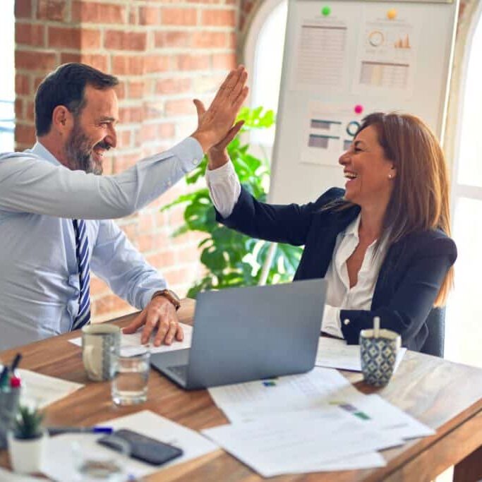 Two business professionals, a man and a woman, sit at a desk smiling and giving each other a high five in an office setting with a laptop, documents, and charts visible.